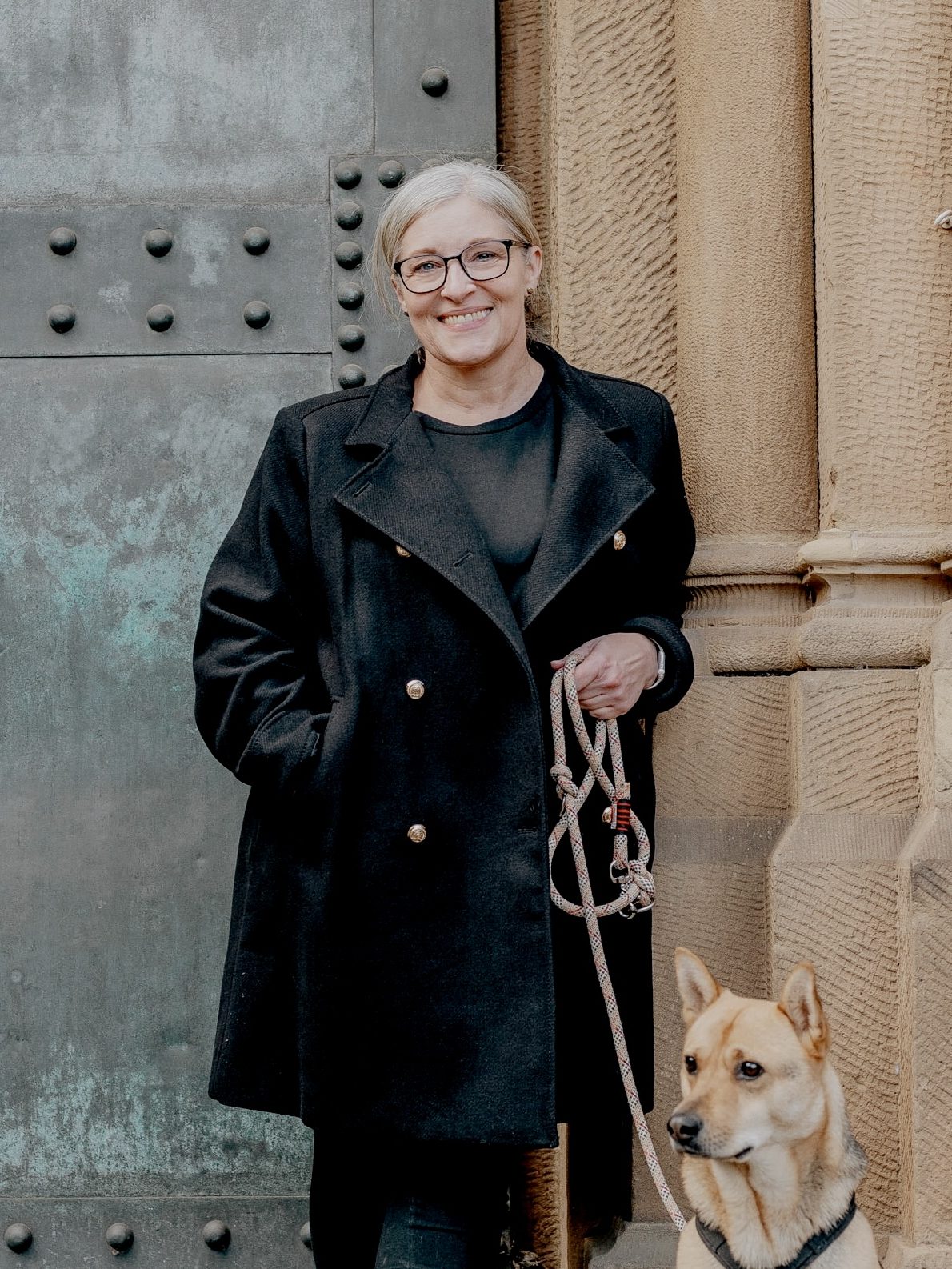 Anja vor derselben Kirchentür, Einstellung ist hier halbnah. Ein Stück des Bauwerks ist nun auch zu sehen. Anja trägt die schwarze Brille und den Mantel mit goldenen Knöpfen, hat in der linken Hand eine Hundeleine und die rechte Hand in der Manteltasche. Der Blick ist freundlich, geht leicht an der Kamera vorbei, der Mund leicht geöffnet zum lächeln. Rechts unten im Bild sind Kopf und Hals vom Mischlingshund Cookie zu sehen. Cookie schaut halb nach links, die Ohren sind aufgestellt. Das Fell hat einen Farbverlauf von Schwarz zu Beige wie bei einem Schäferhund. 